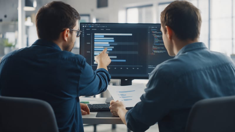 Dos personas de un equipo de desarrollo, de espaldas, revisan en un monitor un gráfico de barras junto a código; una señala la pantalla y la otra sostiene una hoja impresa, con luz de día en una oficina