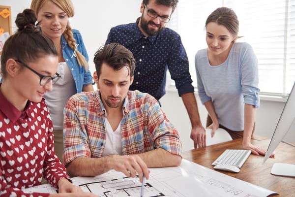 Equipo de desarrollo colaborando en oficina con planos de arquitectura y código en pantallas - instrucciones compartidas para IA