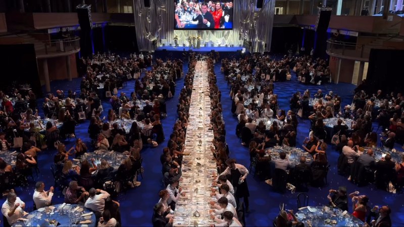 Sala del Palau de Fires de Girona durante la VI Nit Bufona, vista desde arriba: mesas llenas a ambos lados de la mesa central y tres presentadores en el escenario
