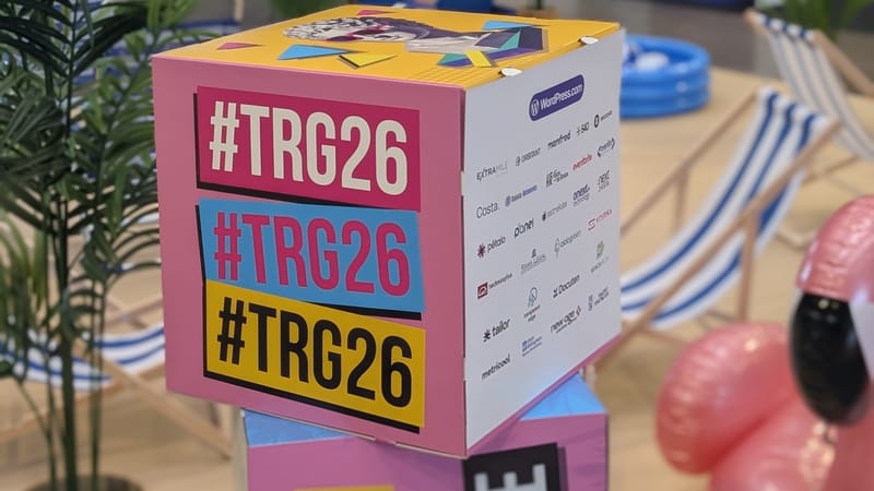 Cubo de cartón con la etiqueta #TRG26 repetida tres veces, en rosa, azul y amarillo, entre plantas y hamacas de una zona común de La Nave